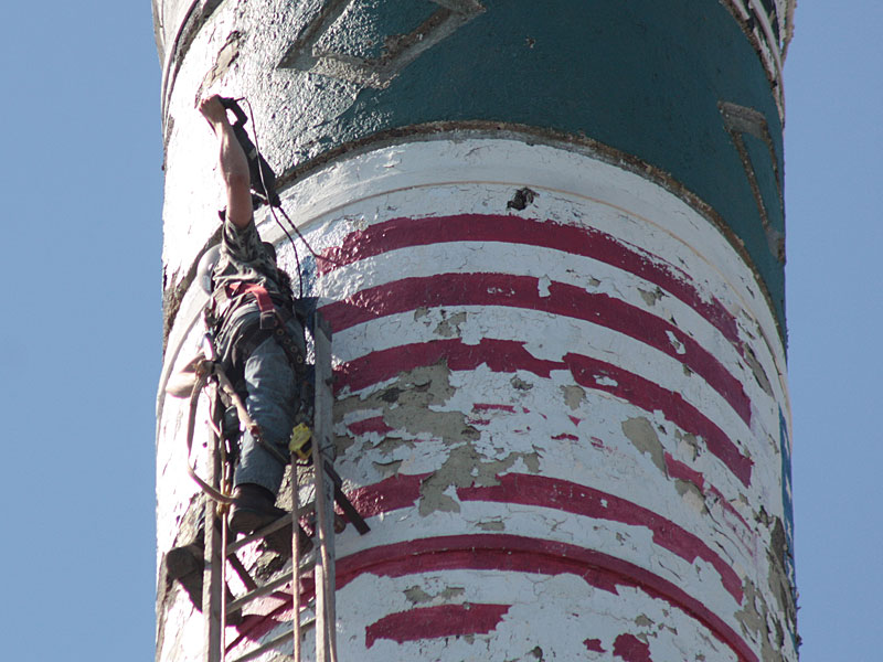 Overhead, and with just one arm, Dustin Frazier uses a lag bolt drill to drill a hole to affix another ladder above the one he is standing on. At this juncture, he is just about at the top of the 170-foot-tall Knox Mill smokestack. (Photo by Holly S. Edwards)