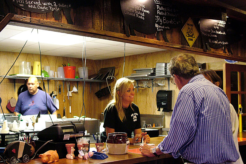 Candace Godwin, daughter of Pig Out owners Ted and Stephanie Guerry, chats with customers below a parade of pig-shaped silhouettes bearing the restaurant's menu. Mel Muriel, left, prepares food in the kitchen. (Photo by Ethan Andrews)