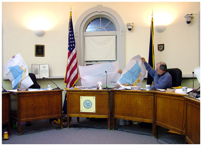 Belfast officials, from left, Councilor Eric Sanders, Mayor Walter Ash, and Councilor Mike Hurley, disappear momentarily behind large maps depicting proposed changes to Front Street Shipyard. (Photo by Ethan Andrews)