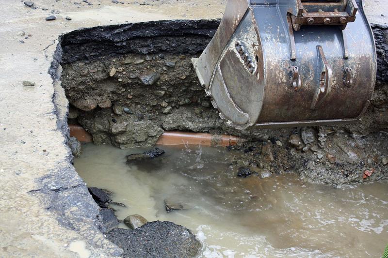 A backhoe picks at the pavement, being careful not to disturb fiber optic lines inside the exposed clay pipe near the broken water pipe. (Photo by Holly S. Edwards)