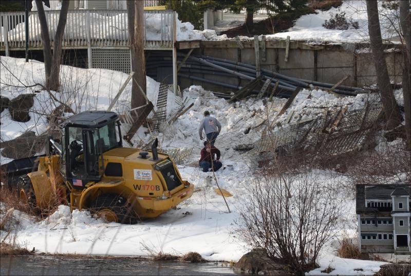Camden Public Works employee Tracy Harford, in burgundy, kneels along the Megunticook River mill pond where the front-end loader he was driving in the parking lot above crashed through the wood sidewalk and a fence and went down the embankment. (Photo by Lynda Clancy)