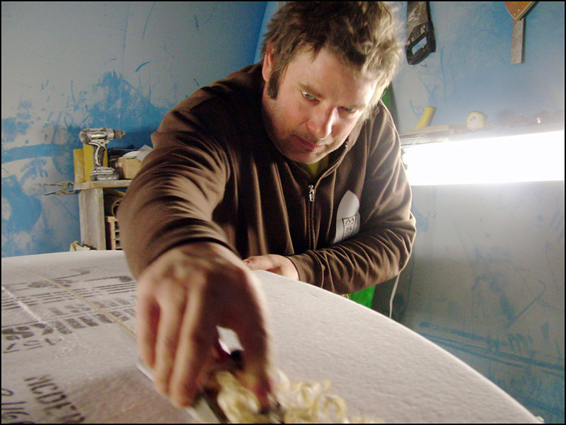 Monroe Surf Co. owner Josh Moody shaves the wooden “stringer” support embedded in the foam “blank” that will eventually become a custom-designed surfboard. For this photo, Moody removed the respirator that he normally wears in the shop. (Photo by Ethan Andrews)