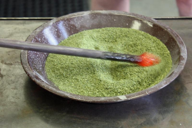 A punty with a glob of 1,800-degree glass on the end is rolled into olive green frit, adhering the color before being reheated, repeated and blended in by rolling on the steel marver table. (Photo by Holly S. Edwards)