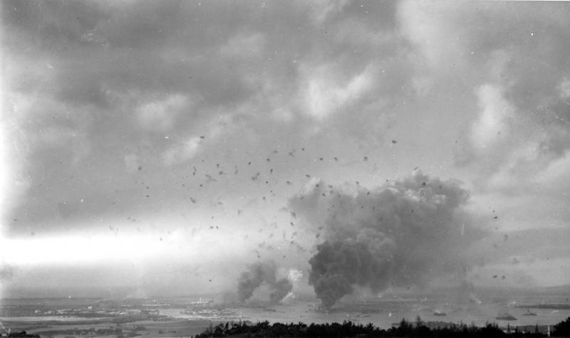 Panorama view of Pearl Harbor, during the Japanese raid on 7 December 1941, with anti-aircraft shell bursts overhead. The photograph looks southwesterly from the hills behind the harbor. Large column of smoke in lower right center is from the burning USS Arizona (BB-39). Smoke somewhat further to the left is from the destroyers Shaw (DD-373), Cassin (DD-372) and Downes (DD-375), in drydocks at the Pearl Harbor Navy Yard.