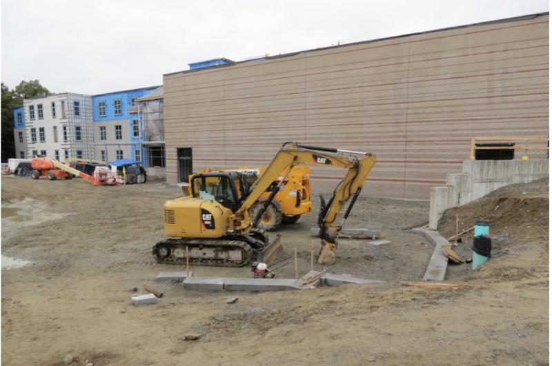 The large window-less (saving money!) wall is the gym with completed exterior siding. To the left is the academic wing. In the space in front of the backhoe and dumptruck is the cement wall that will become the climbing wall and some of the granite edging of the playground has been installed in the center. )Photo courtesy Tom McNealy)