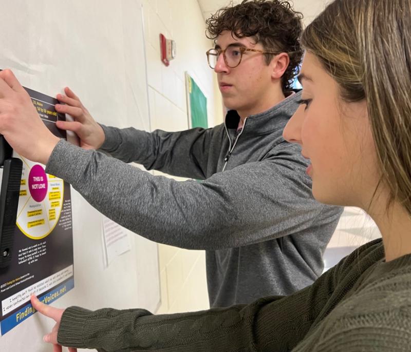 Seniors Sydney Stone, 17, and Owen Berez, 18, put up a This is Not Love poster at Camden Hills Regional High School. The students got more than 25 of the posters up in hallways, the library and cafeteria, and smaller versions of the posters are also going into every bathroom stall in the school. (Photo by Patrisha McLean)