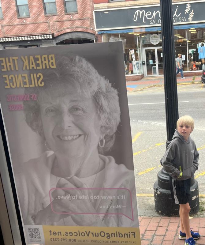 Young passerby takes in Mary Lou’s message from her Finding Our Voices poster at Joe’s Flat Iron Cafe in Skowhegan. Thousands of these posters featuring 45 Maine survivors of domestic abuse are in business windows and also public bathrooms across Maine. (Photo by Patrisha McLean)