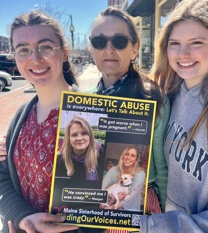 L-r: Lizzie Dunn, Melanie C., and Eva Harvie of Morning Glory Natural Foods in Brunswick on the sidewalk where the Finding Our Voices “Breaking the Silence of Domestic Abuse” march will take place the morning of Saturday, May 10. Folks are encouraged to wear yellow and bring signs expressing what they want the public to know about domestic abuse. The Finding Our Voices poster in the photo is part of a public awareness campaign featuring the photo portraits and quotes of 50 Maine survivors, in business windows of more than 100 downtowns. (Photo by Patrisha McLean) L-r: Lizzie Dunn, Melanie C., and Eva Harvie of Morning Glory Natural Foods in Brunswick on the sidewalk where the Finding Our Voices “Breaking the Silence of Domestic Abuse” march will take place the morning of Saturday, May 10. Folks are encouraged to wear yellow and bring signs expressing what they want the public to know about domestic abuse. The Finding Our Voices poster in the photo is part of a public awareness campaign featuring the photo portraits and quotes of 50 Maine survivors, in business wind