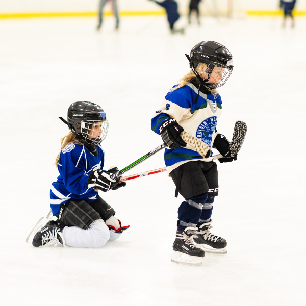Two young kids are having fun learning to skate on ice, while one leads the other around with their hockey sticks.