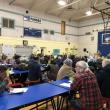a group of people in a school gymnasium, some raising green papers to signify their votes