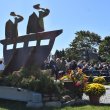 View of a memorial sculpture with ceremony attendees in the background.
