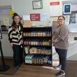 Two women stand next to shelving unit stocked with food