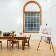 The boardroom at Watts Hall as viewed from the entrance. The room is brightly lit, with white walls, a wood floor, and a large wooden table with vases of fresh cut tulips. There is a large window with an arched top; it's dark outside. There is an easel in the corner, displaying an artist's rendering of what the room might look like filled with people mingling during an event. Photo credit: Nicole Langille Imagery.