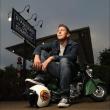 Comedian Lee Hardin sits on a miniature green motor cycle with one foot propped up on a helmet. Behind him his a sign reading "Standard Oil Company".