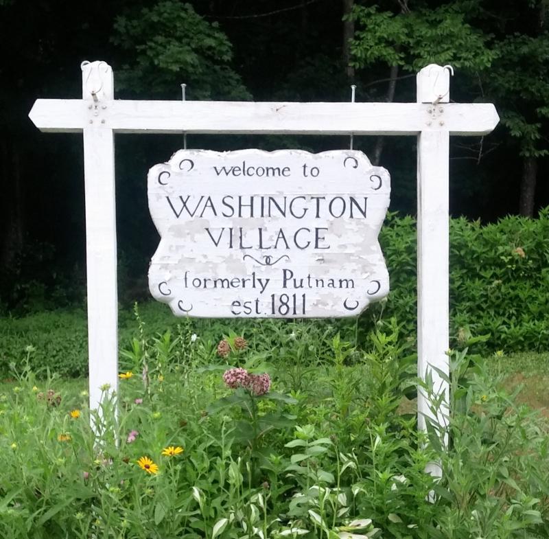 White sign board reading Welcome to Washington Village formerly Putnam est. 1811, with green plants growing around 