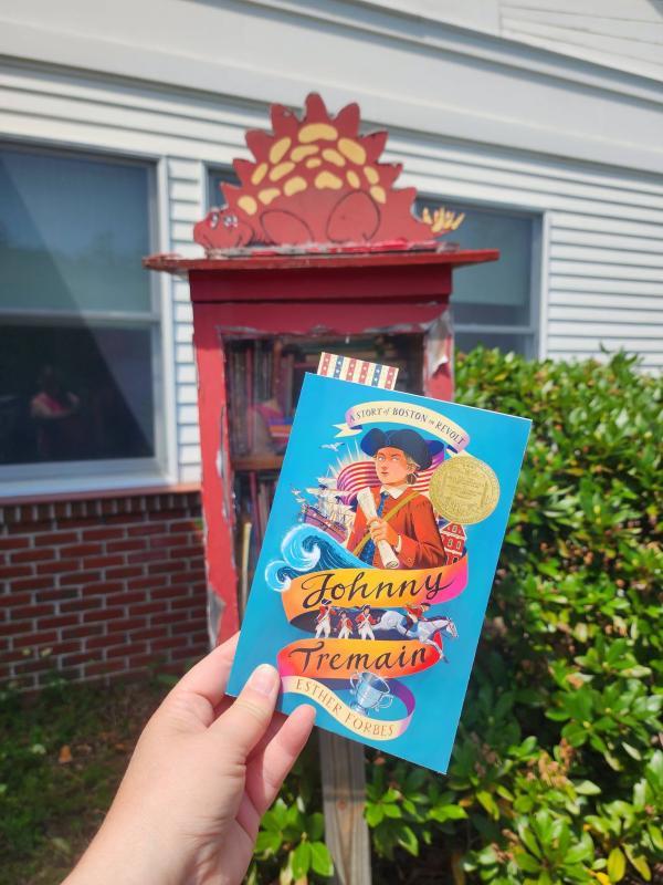 hand holds copy of book in front of little library