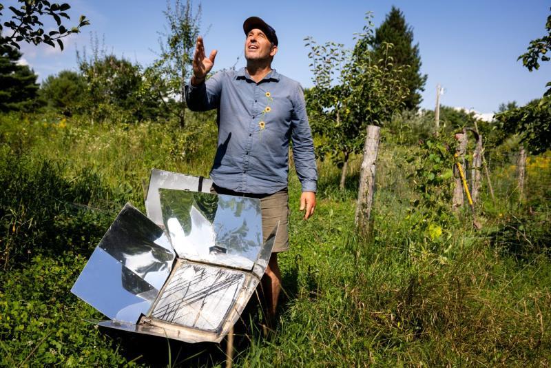 a man stands behind a solar oven in a field.