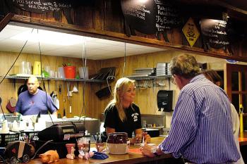 Candace Godwin, daughter of Pig Out owners Ted and Stephanie Guerry, chats with customers below a parade of pig-shaped silhouettes bearing the restaurant's menu. Mel Muriel, left, prepares food in the kitchen. (Photo by Ethan Andrews)