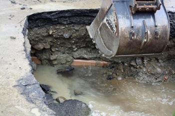 A backhoe picks at the pavement, being careful not to disturb fiber optic lines inside the exposed clay pipe near the broken water pipe. (Photo by Holly S. Edwards)