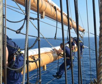 On the yardarm, furling sails. (Photo by John Devine)