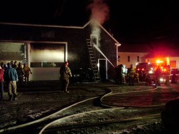 Firefighters pump water into the smoldering upper floor of a commercial building on Swan Lake Avenue in Monroe, Saturday night, while others inspect a garage at left. (Photo by Ethan Andrews)