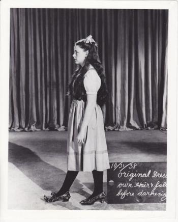 In 1938, Judy models a different dress and wig, and wears one of her earlier ruby slippers on her left foot while displaying a new, ornate design for the shoes on her right foot. (Photo courtesy the collection of John Fricke)