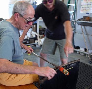 David Jacobson instructs Richard Anderson on how to apply and wrap the thin rod of colored glass, which will become a design element, around the clear glass. (Photo by Holly S. Edwards)