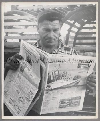 Man in cap and plaid shirt, posing in front of crates with an issue of National Fisherman.
"Oliver Mahonen, a Rockland, Maine, lobster fisherman, looks over the first issue of the National Fisherman combined w/ MCF. Mahonen has been reading MCF almost from the 1st issue and especially likes the classy ads. He says he's pleased w/the expanded coverage offered by the combined pubs. His 28' boat was built in Friendship, ME." No attribution; if you have information about the photographer, please contact Penobscot Marine Museum. (207) 548-2529.)