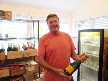 Chef Michel Hetuin co-owner of Chez Michel Restaurant and its newly converted Beach Inn Market, displays one of his favorite champagnes, Veuve Clicquot. Behind him is their collection of wines for sale. Photo by Kay Stephens