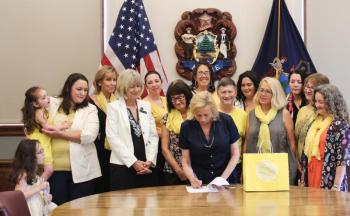Finding Our Voices survivors and supporters flank Governor Janet T. Mills as she ceremoniously signs a bill initiated by the grassroots nonprofit into law. From left to right, Adeline and Eva with mom Nicole LeProhon, Jeannine Lauber Oren, Rep. Vicki Doudera, Governor Mills, Eliza Conley Lepene, Sandra Goulet, Dezarae Caron, Dana Ward, Meg Libby, Finding Our Voices founder and president Patrisha McLean, Kerrie McAnulty, Mary Kamradt, and Jennifer Greensmith. (Photo by Jessi Tracy)