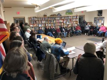 Near a 100 people tried to pack into the Bryant Room for the retail store hearing. (Sarah Masters photo) A group of people stand and sit in a meeting room.