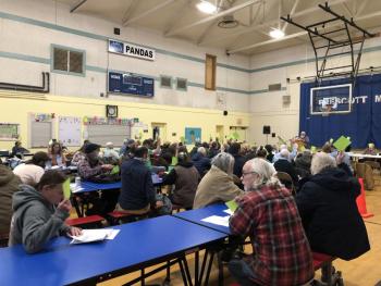 a group of people in a school gymnasium, some raising green papers to signify their votes