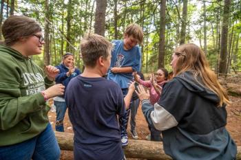 One student tries to balance as they walk across the log without holding onto a rope, although one is made available when needed, while their classmates stand ready to catch them or steady them if they lose their balance during a trust exercise at the 4-H Camp and Learning Center at Tanglewood in Lincolnville. )Photo courtesy of University of Maine Cooperative Extension)
c