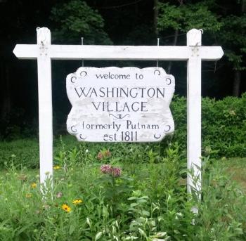 White sign board reading Welcome to Washington Village formerly Putnam est. 1811, with green plants growing around 