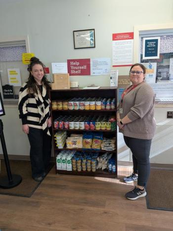 Two women stand next to shelving unit stocked with food