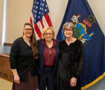 three women pose in front of state, federal flags