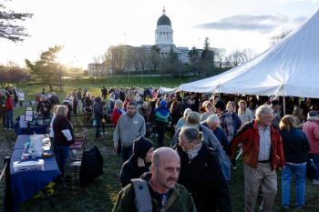 people gather on a lawn with the state house visible in the background