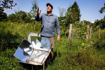 a man stands behind a solar oven in a field.
