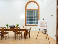 The boardroom at Watts Hall as viewed from the entrance. The room is brightly lit, with white walls, a wood floor, and a large wooden table with vases of fresh cut tulips. There is a large window with an arched top; it's dark outside. There is an easel in the corner, displaying an artist's rendering of what the room might look like filled with people mingling during an event. Photo credit: Nicole Langille Imagery.