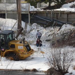 Camden Public Works employee Tracy Harford, in burgundy, kneels along the Megunticook River mill pond where the front-end loader he was driving in the parking lot above crashed through the wood sidewalk and a fence and went down the embankment. (Photo by Lynda Clancy)