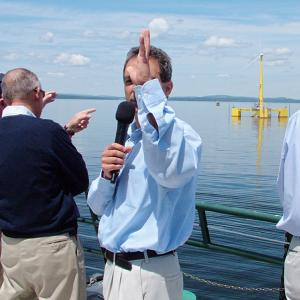 Dr. Habib Dagher, center, of the University of Maine's Advanced Structures & Composites Center, speaks during press conference Thursday marking the ceremonial connection of the first offshore wind turbine in the United States — a 65 foot tall, 1:8 scale prototype, background — to the mainland electrical grid. (Photo by Ethan Andrews)