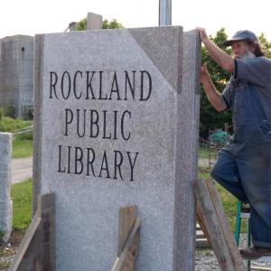 Sculptor Joe Auciello puts the finishing touches on a sculptural sign that was commissioned by the Friends of Rockland Public Library. Carving of the large granite slab took place over the course of the spring on the grounds of the Sail Power and Steam Museum at Sharp's Point South. The work was installed at the corner of Beech and Union streets Tuesday, June 16. (Photo by TimO)