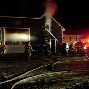 Firefighters pump water into the smoldering upper floor of a commercial building on Swan Lake Avenue in Monroe, Saturday night, while others inspect a garage at left. (Photo by Ethan Andrews)
