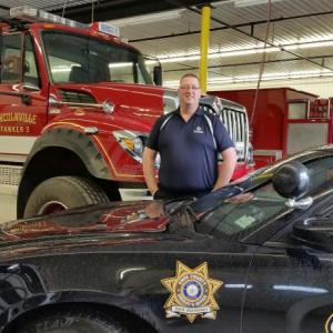 Justin Hills, wearing his Northeast Health uniform, stands with a Lincolnville fire truck and Knox County Sheriff cruiser, two of the four vehicles he drives as a first responder.