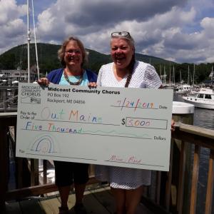 With Camden Harbor in the backgroud, Mim Bird, the chair of MCCs board, presents a check to Jeanne Dooley, director of OUT Maine. (Photo by Midcoast Community Chorus)