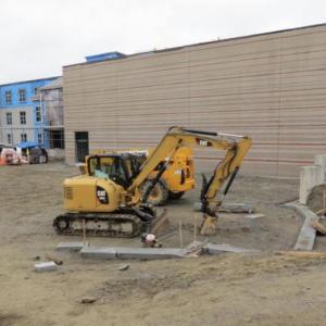 The large window-less (saving money!) wall is the gym with completed exterior siding. To the left is the academic wing. In the space in front of the backhoe and dumptruck is the cement wall that will become the climbing wall and some of the granite edging of the playground has been installed in the center. )Photo courtesy Tom McNealy)