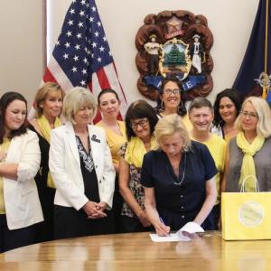 Finding Our Voices survivors and supporters flank Governor Janet T. Mills as she ceremoniously signs a bill initiated by the grassroots nonprofit into law. From left to right, Adeline and Eva with mom Nicole LeProhon, Jeannine Lauber Oren, Rep. Vicki Doudera, Governor Mills, Eliza Conley Lepene, Sandra Goulet, Dezarae Caron, Dana Ward, Meg Libby, Finding Our Voices founder and president Patrisha McLean, Kerrie McAnulty, Mary Kamradt, and Jennifer Greensmith. (Photo by Jessi Tracy)