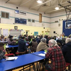 a group of people in a school gymnasium, some raising green papers to signify their votes