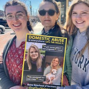 L-r: Lizzie Dunn, Melanie C., and Eva Harvie of Morning Glory Natural Foods in Brunswick on the sidewalk where the Finding Our Voices “Breaking the Silence of Domestic Abuse” march will take place the morning of Saturday, May 10. Folks are encouraged to wear yellow and bring signs expressing what they want the public to know about domestic abuse. The Finding Our Voices poster in the photo is part of a public awareness campaign featuring the photo portraits and quotes of 50 Maine survivors,  in business wind