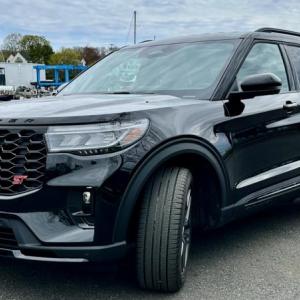The red ST badge and black grille denote the 400-horsepower performance variant of the 2025 Ford Explorer; other visual clues include red brake calipers and these blacked-out 21-inch aluminum wheels. The view out over the high, squared-off nose is distinctly pickup-truck. This color is Agate black Metallic. 